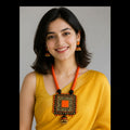 A woman with dark shoulder-length hair wearing a bright yellow-orange statement necklace with a square patterned pendant and matching orange-and-gold earrings, paired with a yellow top, against a plain gray background.