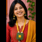 A woman with long black hair smiling, wearing a red embroidered kurta, a mustard yellow dupatta, and a large green and yellow geometric pendant necklace with matching earrings, set against a pink wall with green leaves.