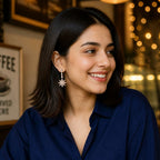 A woman with dark shoulder-length hair smiling in a cafe setting, wearing a navy blue button-up shirt and light blue jeans, with a coffee cup in the foreground and warm bokeh lights in the background.