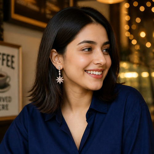 A woman with dark shoulder-length hair smiling in a cafe setting, wearing a navy blue button-up shirt and light blue jeans, with a coffee cup in the foreground and warm bokeh lights in the background.
