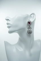 A pair of silver-toned dangle earrings with round red gemstone tops and sparkling clear pavé teardrop drops, displayed in a black velvet jewelry box on a patterned orange and beige fabric surface.