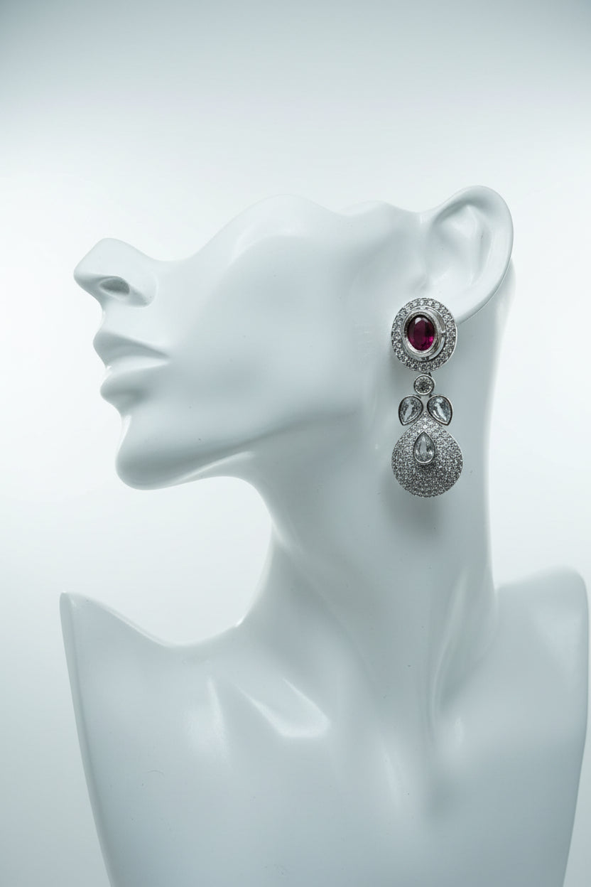 A pair of silver-toned dangle earrings with round red gemstone tops and sparkling clear pavé teardrop drops, displayed in a black velvet jewelry box on a patterned orange and beige fabric surface.