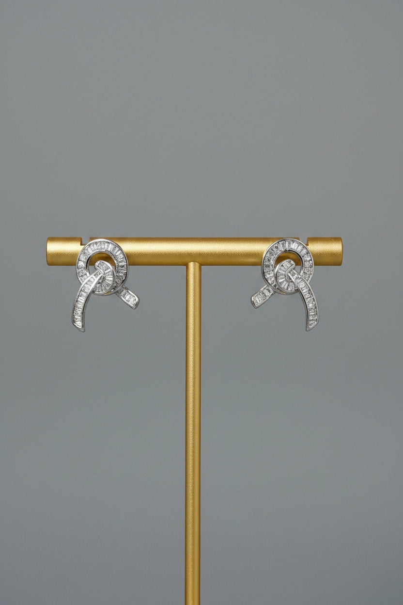 A pair of silver-toned hoop earrings with clear stones, displayed on a white Pacific brand card mounted on a small wooden stand, set against an orange and beige fabric background.