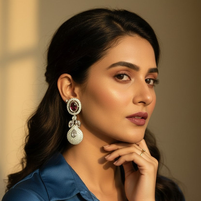 A woman with dark hair wearing a blue satin shirt and a large silver-toned dangle earring with a red stone, posing against a warm beige background.