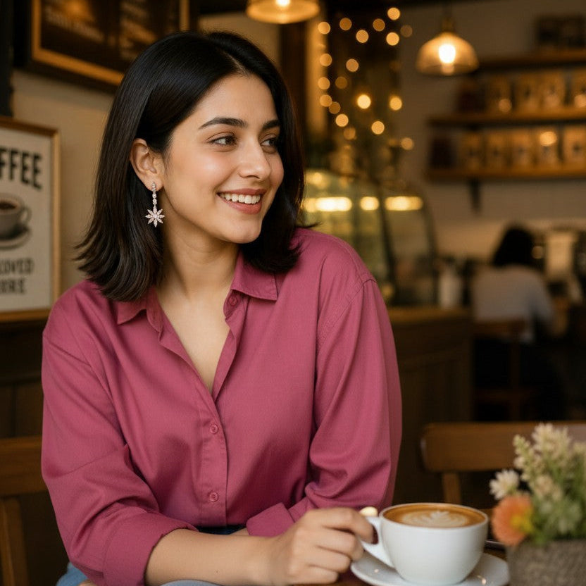 A woman with dark shoulder-length hair sits at a cafe table wearing a dusty rose pink button-up shirt and light blue jeans, holding a white coffee cup. The background shows a warm, softly blurred cafe interior with string lights and wooden furniture.