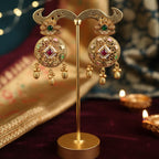 A pair of ornate gold-toned Indian jhumka earrings with green and pink stones, displayed on a gold stand against a dark, festive background with string lights and a sari.