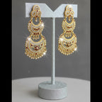 A pair of ornate gold-toned chandelier earrings with red and white stone detailing, displayed on a glittery silver stand against a gray background.