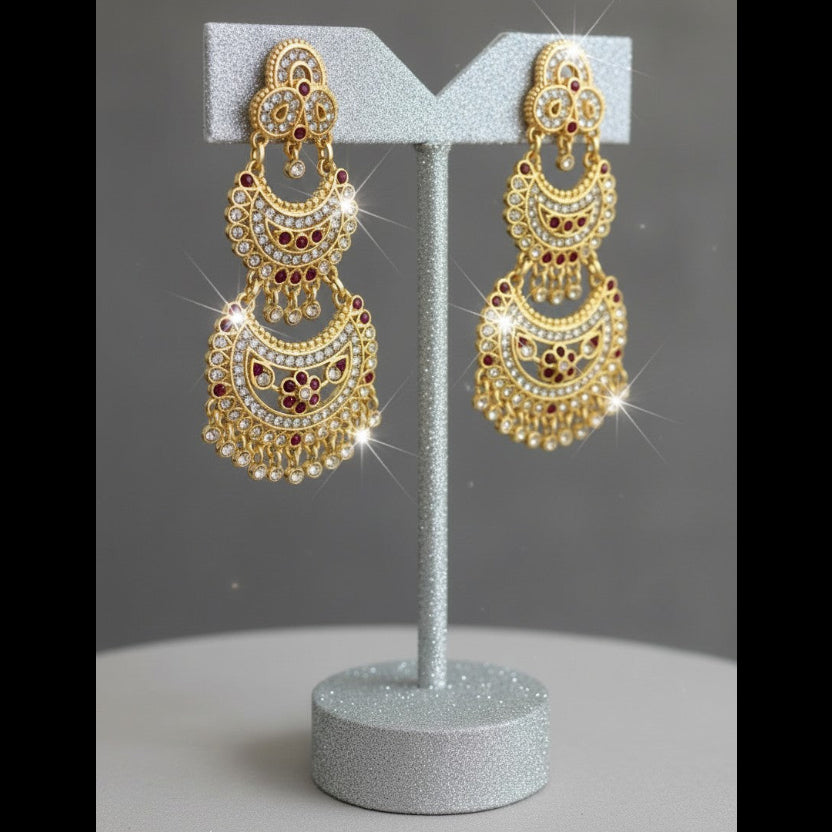 A pair of ornate gold-toned chandelier earrings with red and white stone detailing, displayed on a glittery silver stand against a gray background.