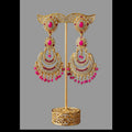 Gold-toned traditional Indian earrings displayed on a stand, featuring crescent-shaped dangle earrings with pink stones, white stones, and dangling bead chains, set against a dark brown background with draped magenta and teal fabric.