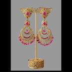Gold-toned traditional Indian earrings displayed on a stand, featuring crescent-shaped dangle earrings with pink stones, white stones, and dangling bead chains, set against a dark brown background with draped magenta and teal fabric.