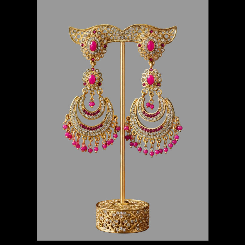 Gold-toned traditional Indian earrings displayed on a stand, featuring crescent-shaped dangle earrings with pink stones, white stones, and dangling bead chains, set against a dark brown background with draped magenta and teal fabric.