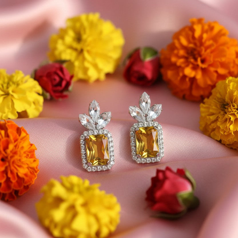 A pair of silver-toned earrings with rectangular yellow gemstones and clear crystal leaf-shaped tops, arranged on pink satin fabric with yellow and orange flowers around them.