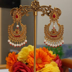 A pair of ornate gold-toned Indian jhumka earrings with red stone accents and dangling white pearl beads, displayed on a decorative gold stand in front of a blurred background with orange, yellow, and red flowers.