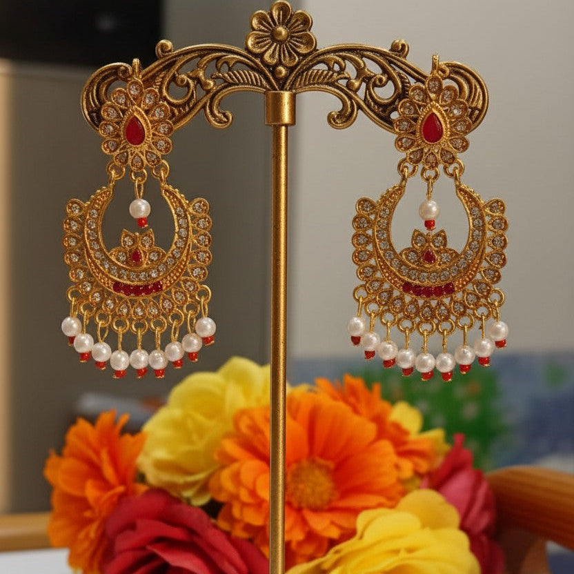 A pair of ornate gold-toned Indian jhumka earrings with red stone accents and dangling white pearl beads, displayed on a decorative gold stand in front of a blurred background with orange, yellow, and red flowers.