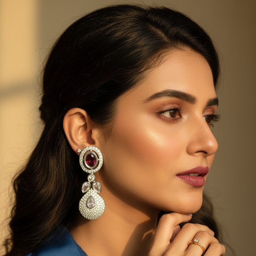 A woman in a blue satin shirt wearing a large, ornate silver-toned dangle earring with a deep red oval stone and clear crystal details, shown in a warm, sunlit portrait.