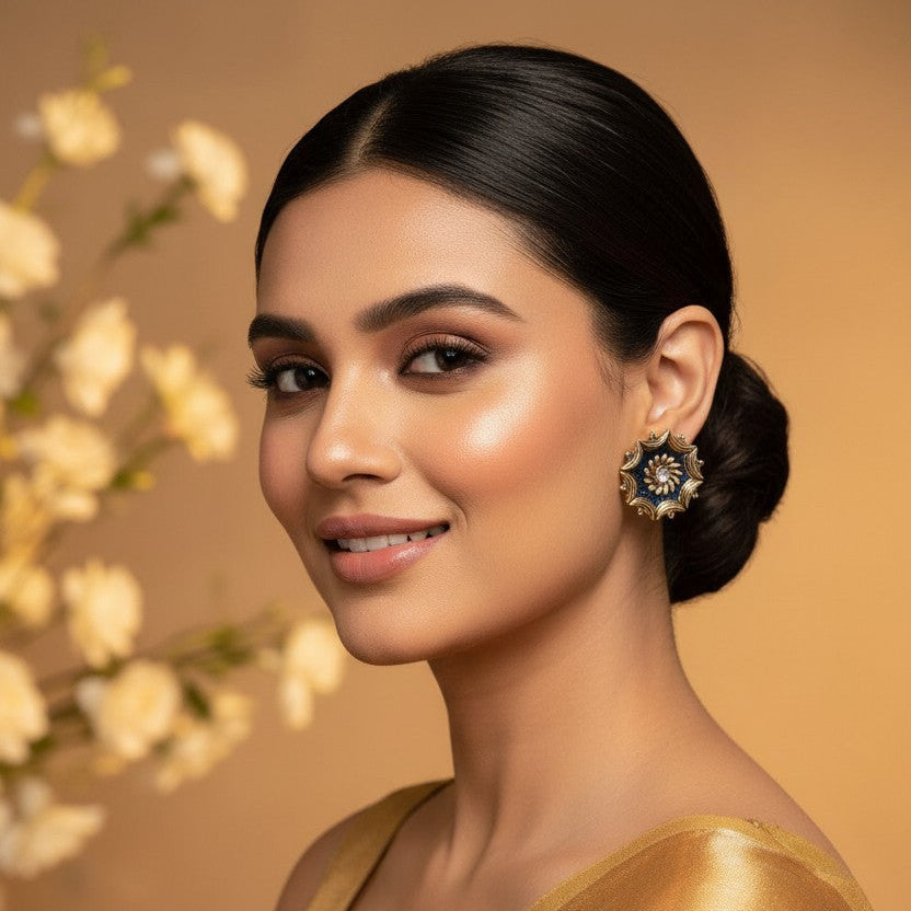 A woman with dark hair styled in a low bun wears a golden yellow embroidered saree and a matching ornate earring, set against a warm beige background with soft white flowers on the left.