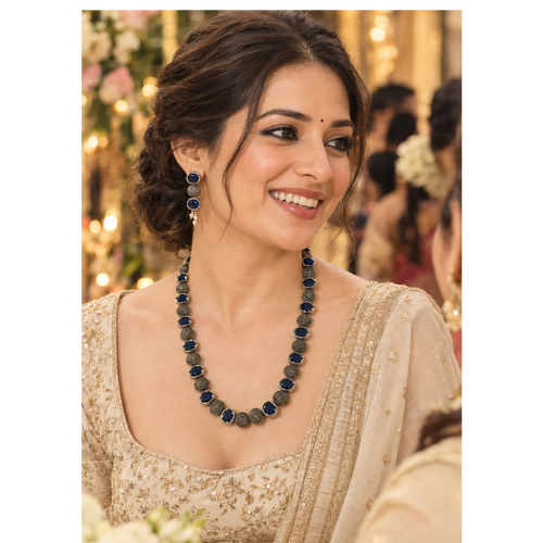 A woman wearing a traditional Indian outfit with a beaded necklace and matching earrings, smiling in a decorated event setting.