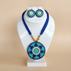 A bohemian-style evil eye jewelry set featuring a round pendant necklace and matching drop earrings, displayed on a white jewelry bust. The jewelry has a vibrant blue cord, gold-toned beads, and circular pendants/earrings decorated with turquoise, white, and orange patterns on a dark blue background.