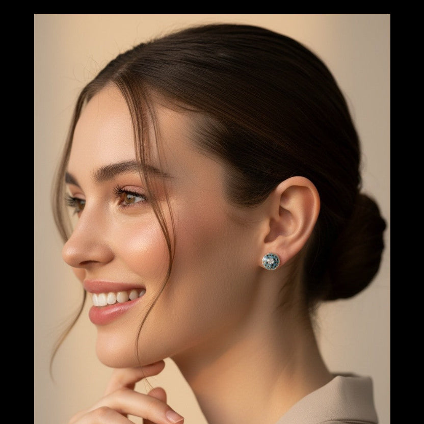 A woman with dark hair in a low bun is shown in profile wearing a small round stud earring with a light blue center and silver-toned setting. She is wearing a beige top against a soft beige background.