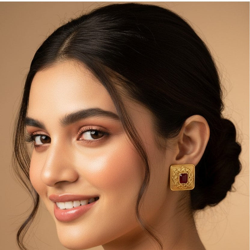 A woman with dark hair in a low bun is shown in profile wearing a square gold earring with a red center stone, against a warm beige background.