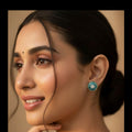 A close-up portrait of a woman wearing a round turquoise and white beaded earring, with her hand near her chin against a warm beige background.