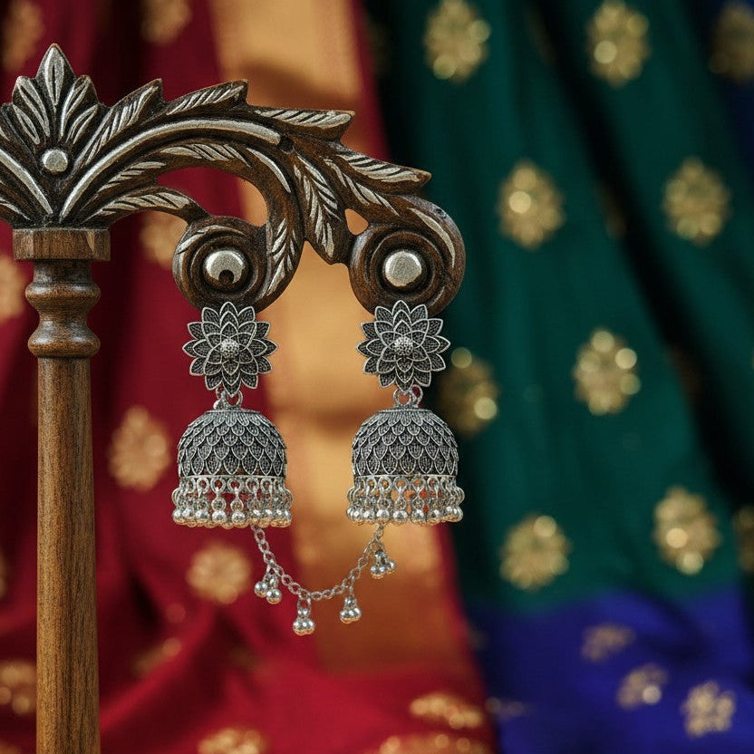 Silver-toned Indian jhumka earrings displayed on a carved wooden stand, with draped red, green, and blue fabric in the background.