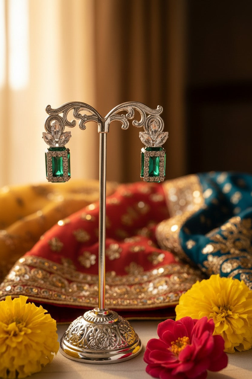 A pair of ornate silver-toned earrings with rectangular green gemstones, displayed on a decorative silver stand. The background is softly blurred with traditional red and blue embroidered fabrics, yellow marigold flowers, and a warm indoor setting.