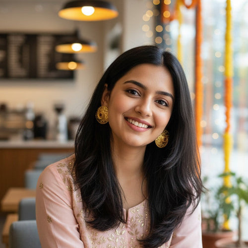 A woman with long black hair is smiling while wearing a light pink traditional outfit with gold embroidery and gold earrings, seated in a cafe setting.