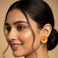 A close-up portrait of a woman wearing a gold-toned traditional flower-shaped earring with a pink stone center, against a warm beige background.