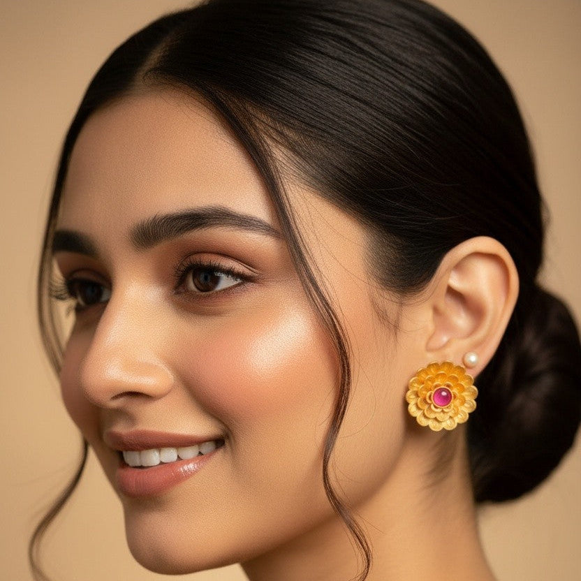A close-up portrait of a woman wearing a gold-toned traditional flower-shaped earring with a pink stone center, against a warm beige background.