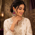 A woman wearing a cream-colored embroidered traditional outfit with gold detailing, styled with silver statement earrings featuring pink stones and pearl bead drops, posed in an ornate indoor setting.