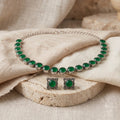 A silver-tone jewelry set featuring a necklace and matching earrings, each adorned with round green stones in ornate square settings, displayed on beige fabric and a stone slab.
