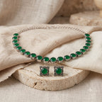 A silver-tone jewelry set featuring a necklace and matching earrings, each adorned with round green stones in ornate square settings, displayed on beige fabric and a stone slab.