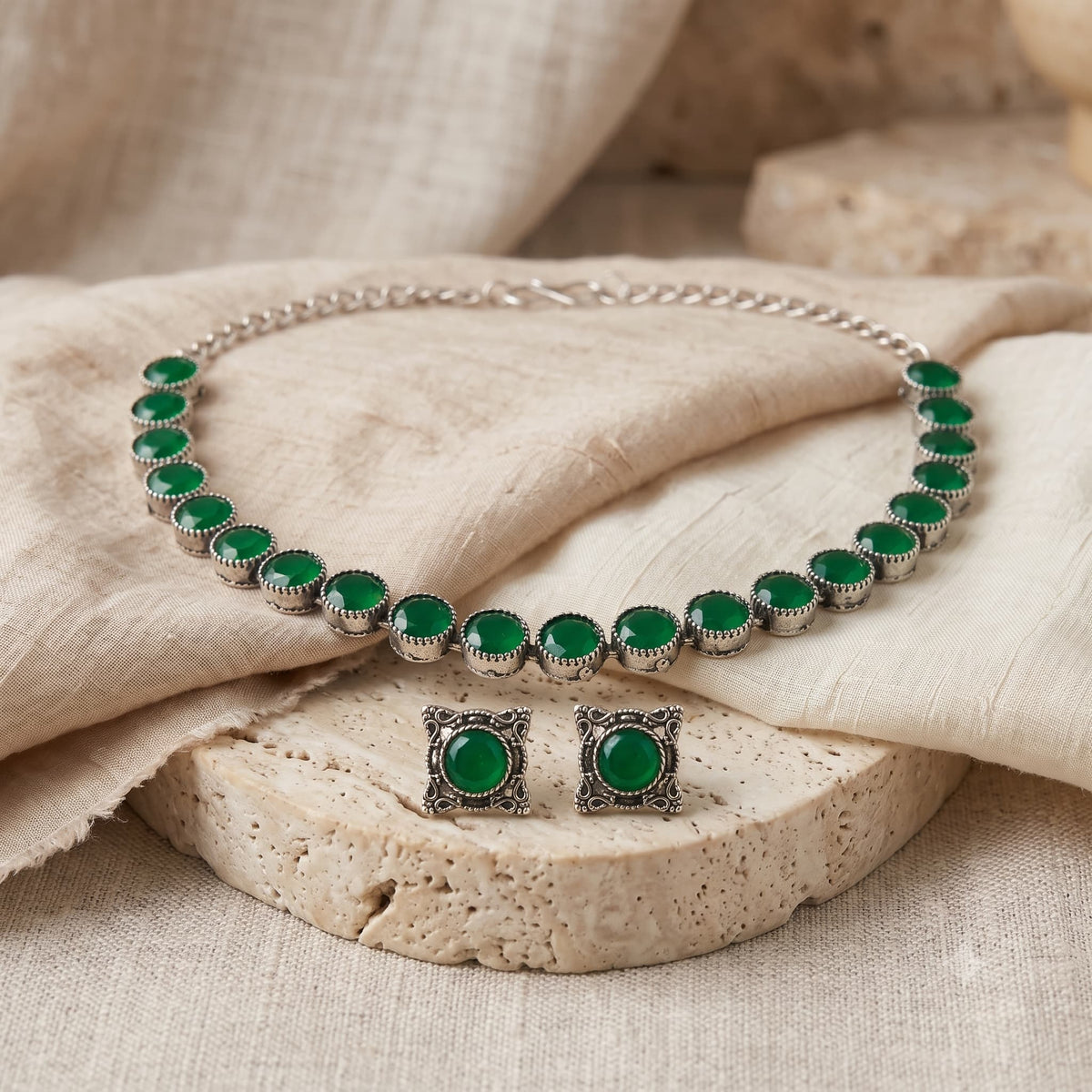 A silver-tone jewelry set featuring a necklace and matching earrings, each adorned with round green stones in ornate square settings, displayed on beige fabric and a stone slab.