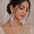 A close-up of a woman wearing ornate silver-toned statement earrings with pink and green stone accents and dangling chain drops, paired with a light beige embellished traditional outfit against a soft cream backdrop.