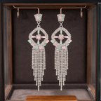 A pair of long dangling statement earrings displayed inside a jewelry box. The earrings have a silver-tone geometric frame set with pale pink stones and mint green accents, with cascading chains of clear rhinestones hanging below.