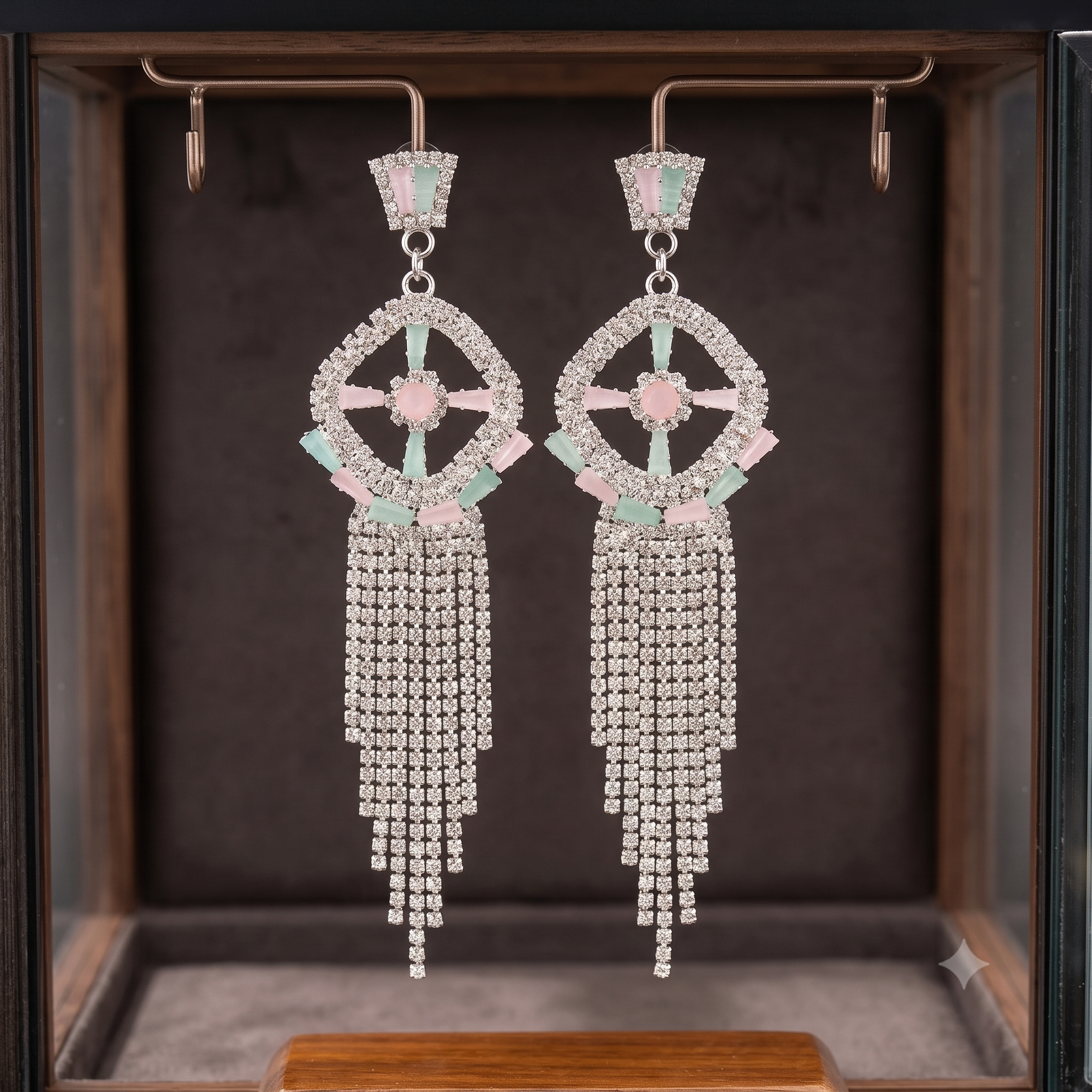 A pair of long dangling statement earrings displayed inside a jewelry box. The earrings have a silver-tone geometric frame set with pale pink stones and mint green accents, with cascading chains of clear rhinestones hanging below.