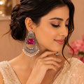 A woman wearing an ornate silver-toned earring with a large pink gemstone and dangling pearl-like accents, photographed in a warm indoor setting.