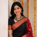 A woman wearing a black and red sari with gold zari embroidery, a matching red blouse, and a gold-toned statement necklace, standing indoors against a light wall with marigold garlands.