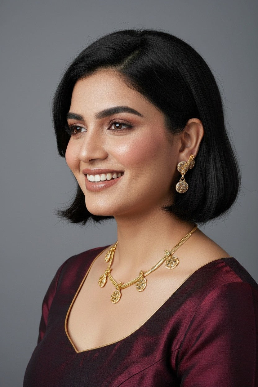A woman with a short black bob hairstyle is shown in a close-up portrait, wearing a gold necklace and matching gold earrings with a deep burgundy satin top against a solid gray background.