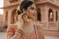 A woman in traditional Indian attire wearing ornate gold-toned earrings and bangles, posed outdoors against a pink sandstone architectural background.