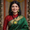 A woman wearing a traditional Indian green and red saree with gold zari borders, a red blouse, and a large round gold pendant necklace, set against a festive background with marigold garlands and warm bokeh lights.