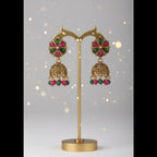 Gold-toned traditional Indian jhumka earrings with green and pink enamel detailing, displayed on a black stand with a white V.S Jewelry card, resting on a newspaper surface.