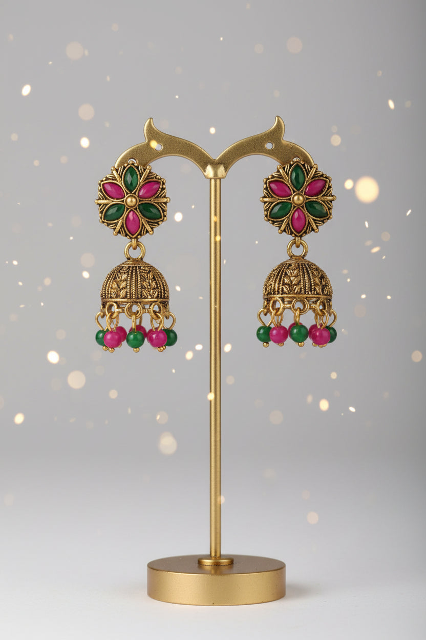 Gold-toned traditional Indian jhumka earrings with green and pink enamel detailing, displayed on a black stand with a white V.S Jewelry card, resting on a newspaper surface.