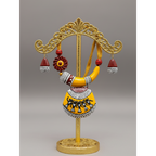 A terracotta clay jewelry set by Avigna Terracotta, featuring a large necklace and matching earrings displayed on a round woven mat. The jewelry is decorated with yellow, red, black, and white patterns, including a curved collar and a large pendant with figures.