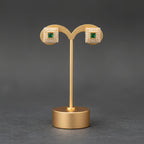 A pair of gold-toned stud earrings with square pavé settings and green square centers, displayed on a gold stand against a dark gray background.