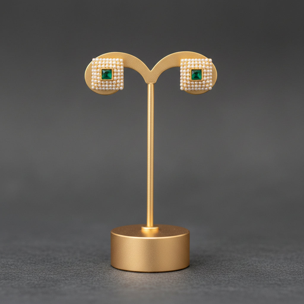 A pair of gold-toned stud earrings with square pavé settings and green square centers, displayed on a gold stand against a dark gray background.