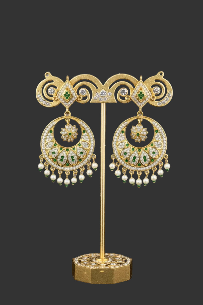 A pair of gold-toned traditional Indian jhumka earrings with intricate filigree work, small green stone accents, and dangling pearl-like beads, shown on a plain light background.