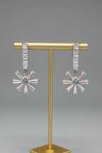 A pair of silver-toned dangle earrings with baguette-cut clear stones, displayed on a gold T-stand against a neutral gray background.