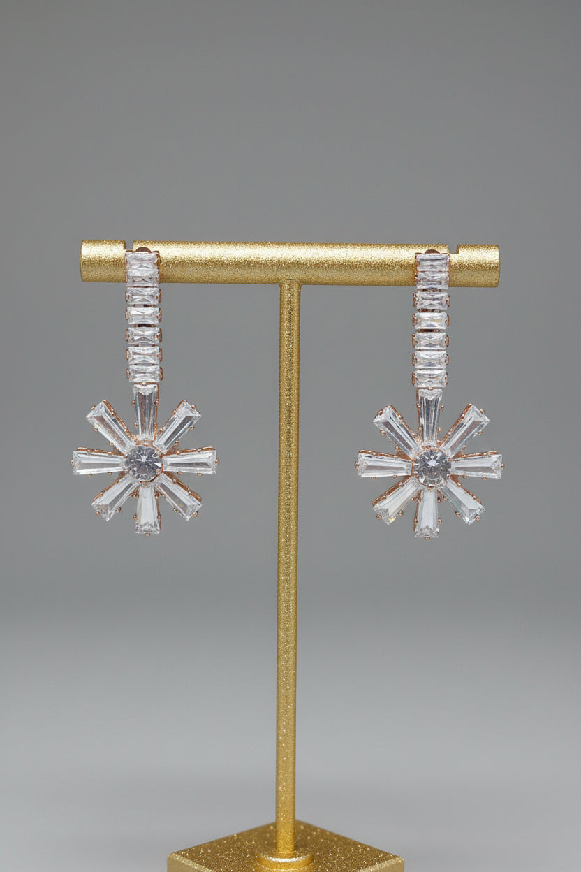 A pair of silver-toned dangle earrings with baguette-cut clear stones, displayed on a gold T-stand against a neutral gray background.